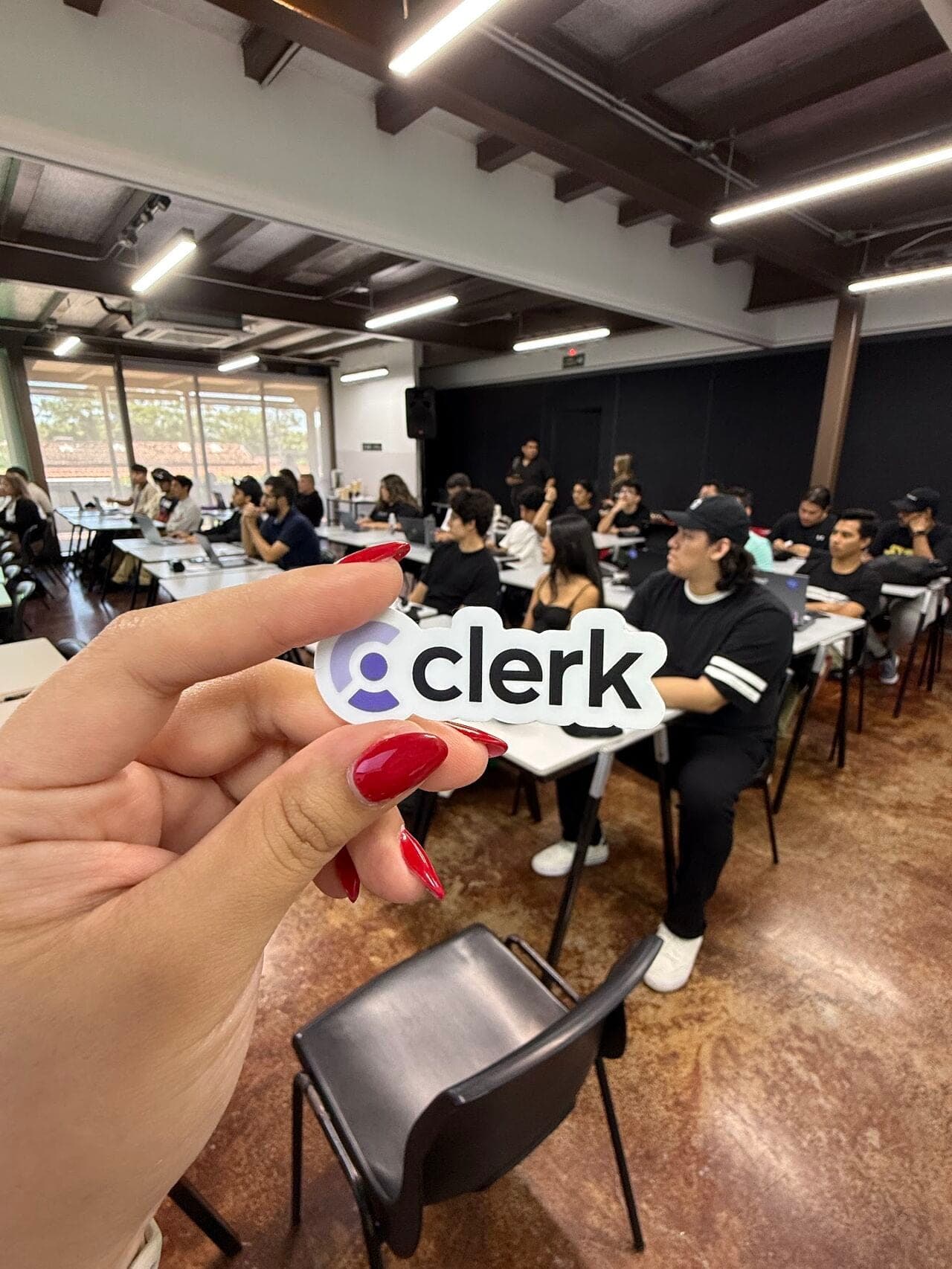 Hand holding a Clerk sticker in front of a full meetup room